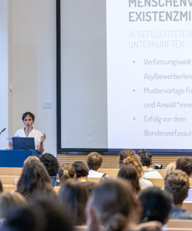 Hörsaal aus Studierendenperspektive, Redner*in Sarah Lincoln am Lehrpult vor einer Powerpoint Präsentation